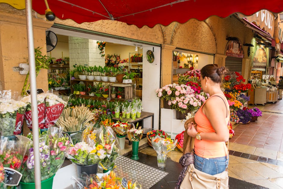 A woman in a peach tank top and denim shorts stands outside a flower shop, selecting a floral gift. The shop offers a variety of fresh flowers including roses, lilies, and carnations in vibrant pinks,