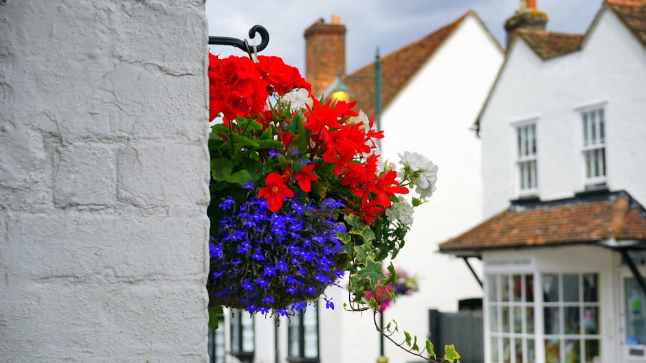 A vibrant hanging flower basket attached to a white brick wall, featuring large red geraniums, white petunias, and clusters of small blue lobelias, with green foliage cascading down. The background sh