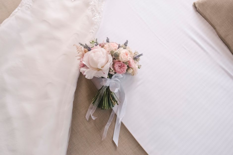 A delicate bridal bouquet featuring soft pink and white roses, peonies, and lavender sprigs, all arranged in a compact, rounded style. The flowers are fresh with gently textured petals and green stems