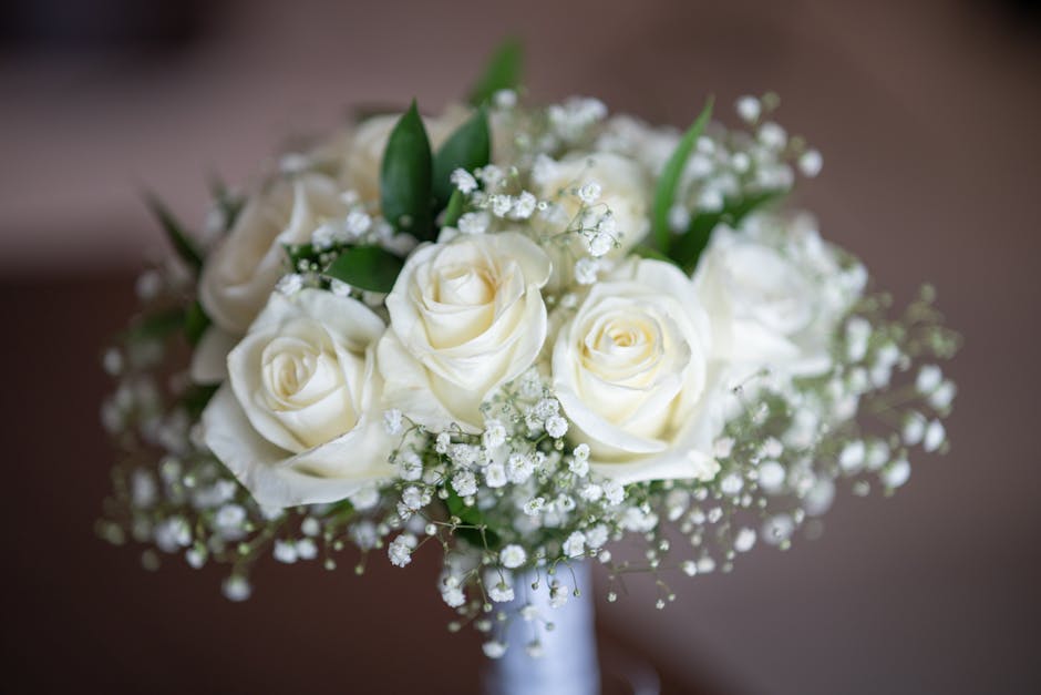 A bouquet of fresh, white roses arranged with small white baby's breath flowers and green foliage, wrapped in delicate white paper, placed on a brown surface. The roses display soft, velvety petals wi