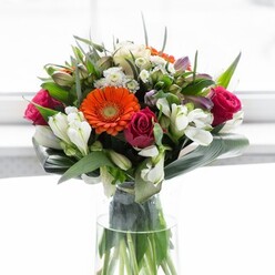 This striking floral bouquet, artfully composed by a seasoned Mottingham florist, embodies both exuberance and gentle sophistication. Creamy peach and bold orange gerbera daisies with dark chocolate centres radiate from the heart of the display, enveloped by plush, magenta roses whose petals unfurl with an air of antique hope. Soft white alstroemeria, like scattered pearls, weave brightness and intricate texture into the assortment, while lighter pink accents and diminutive white filler blooms whisper of springtime along Mottingham's leafy paths. The arrangement's lush greenery-a blend of broad aspidistra leaves expertly curved into a protective, sculptural collar and clusters of sharper, vivid foliage-anchors the bouquet with depth and natural grace. The stems are visible through the tall, clear glass vase, capturing the sense of freshness and authenticity found in renowned Mottingham flower shops. Bathed in diffused, luminous light, each colour and petal becomes more vibrant, while the backdrop remains minimal, drawing all eyes to the bouquet's artistic unity. There's a subtle fragrance-a melody of garden rose, green leaf, and delicate lily-that brings the senses to life. This arrangement effortlessly suits a celebratory Sunday brunch, a heartfelt thank you, or the start of a new home near Elmstead Woods. Every element of this piece feels considered and symbolic-an invitation to optimism and gratitude woven through Mottingham's local spirit.